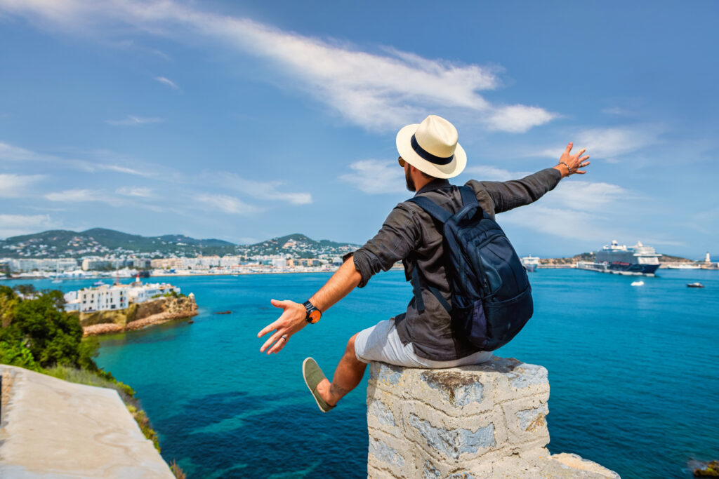 Man with a backpack and straw hat sitting on a stone wall, overlooking a vibrant blue sea and coastal city, embodying adventure and travel experiences with Tropical Travel Adventures in Sacramento.