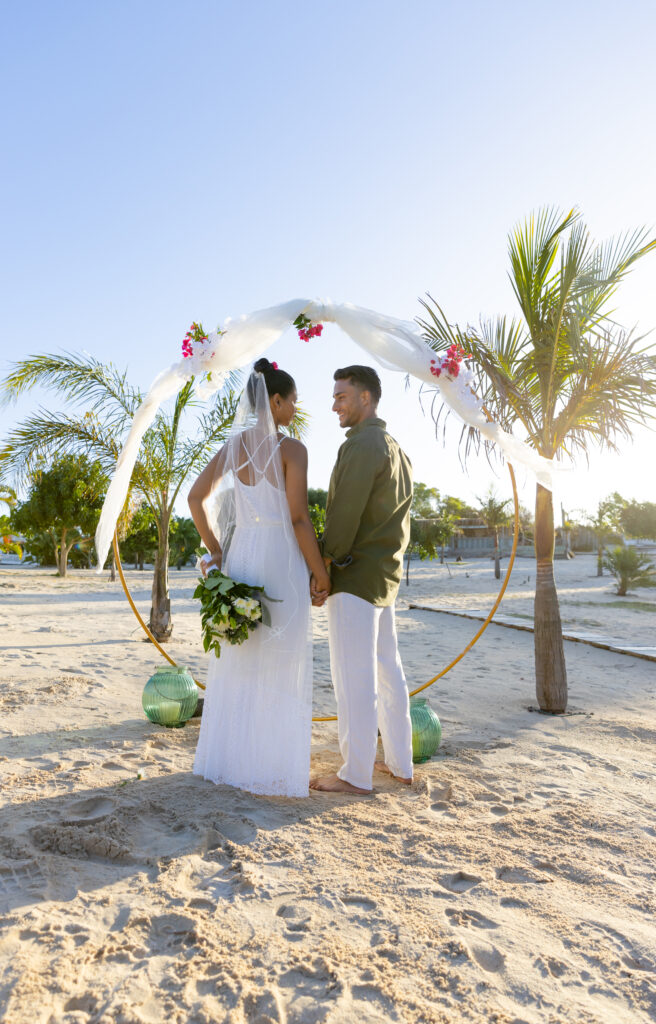 Couple holding hands in a beach wedding setting, standing under a floral arch with palm trees and sand, representing an inclusive destination wedding vacation.