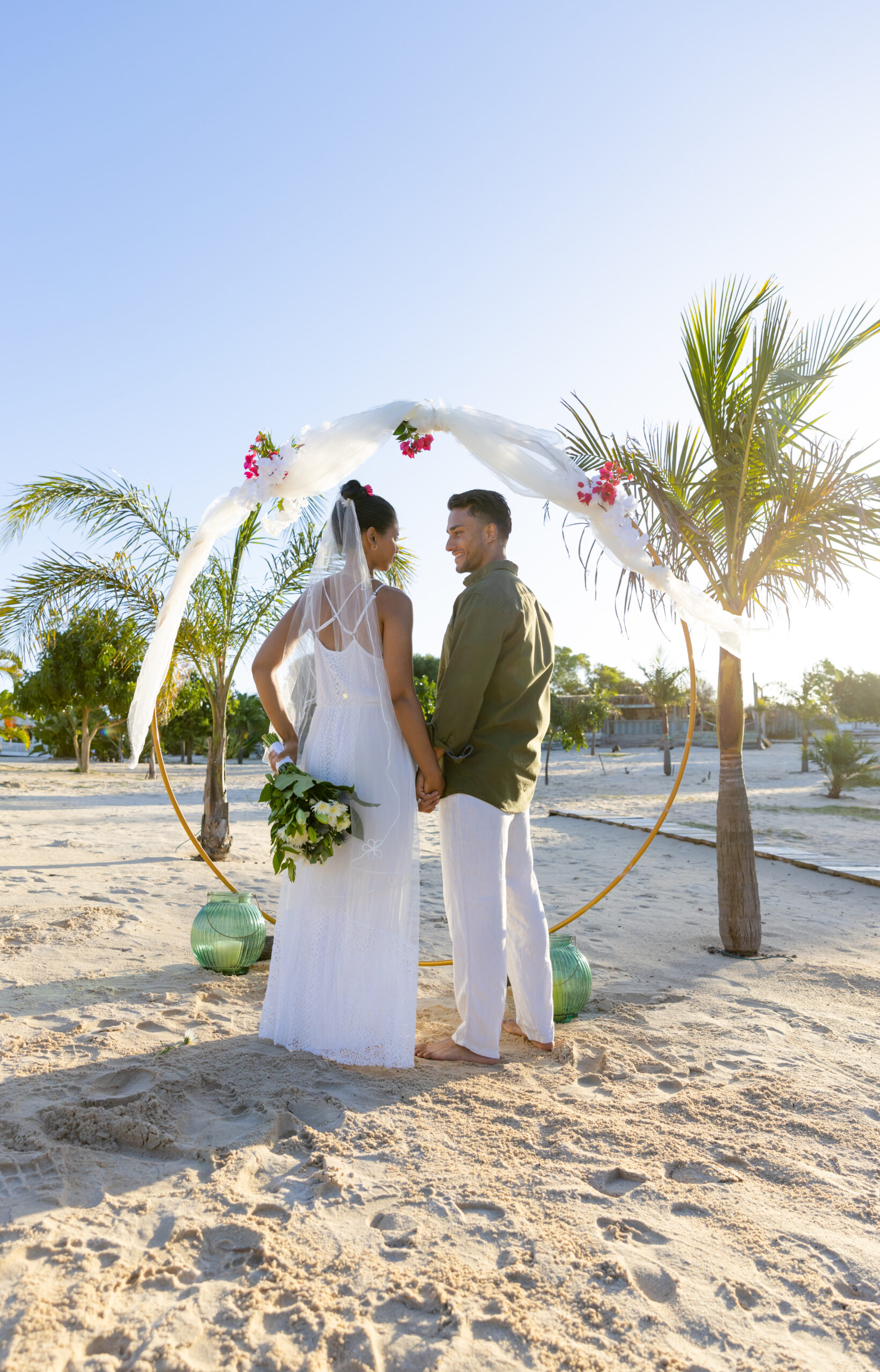 Couple holding hands under a floral wedding arch on a beach, symbolizing inclusive destination wedding vacations.