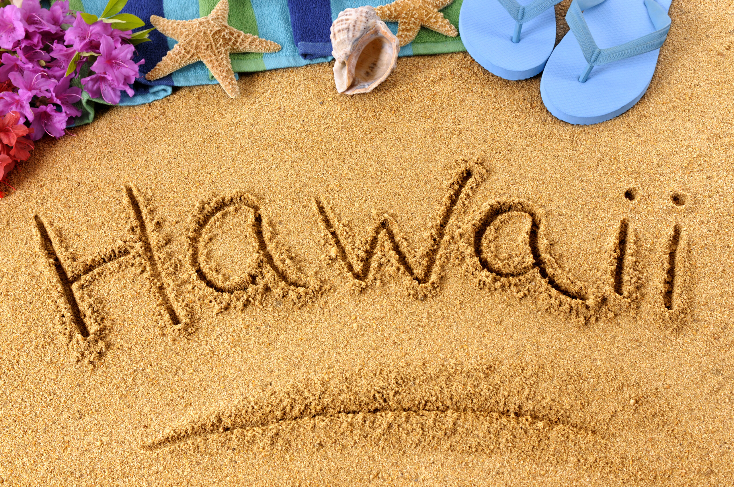 Hawaii written in sand with colorful flowers, a starfish, a seashell, and blue flip-flops, symbolizing adventure travel in Hawaii.