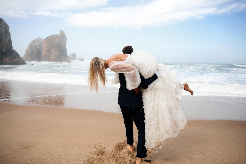 Couple celebrating a destination wedding on a beach, groom carrying bride in a wedding dress, ocean waves and rocky cliffs in the background, emphasizing romantic all-inclusive wedding travel experiences.