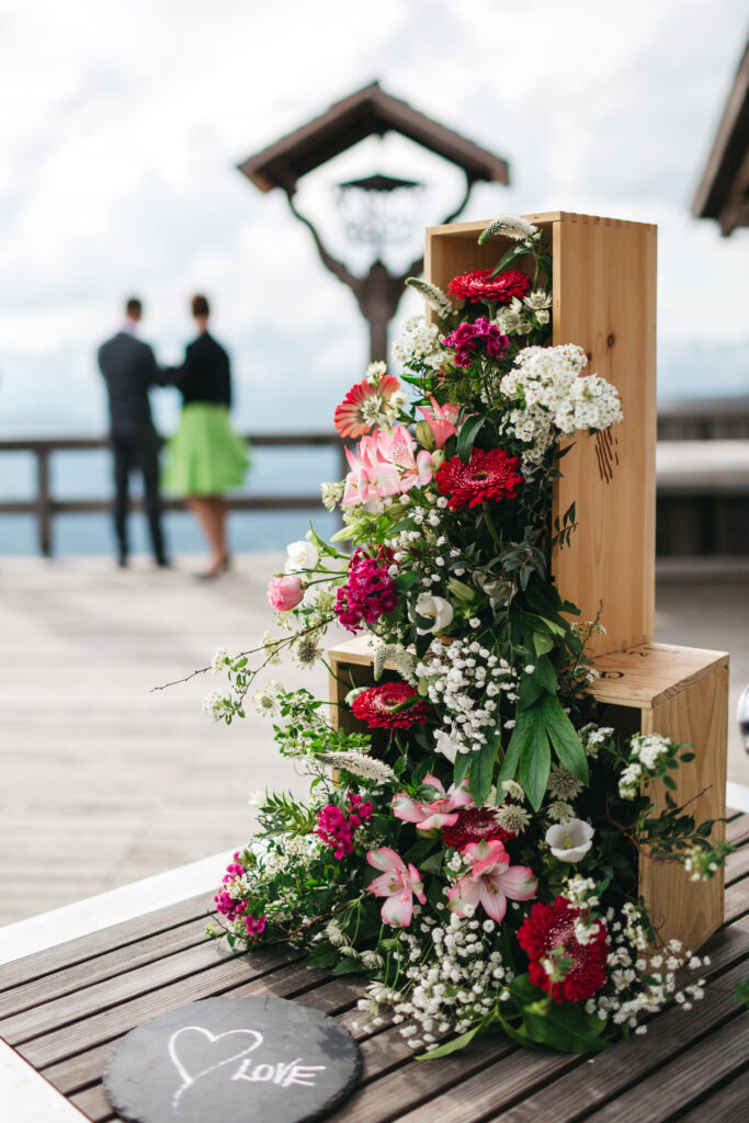 Wooden blocks adorned with vibrant flowers, featuring a slate with "LOVE," in the foreground; a couple in wedding attire stands in the background, highlighting a romantic destination wedding setting.