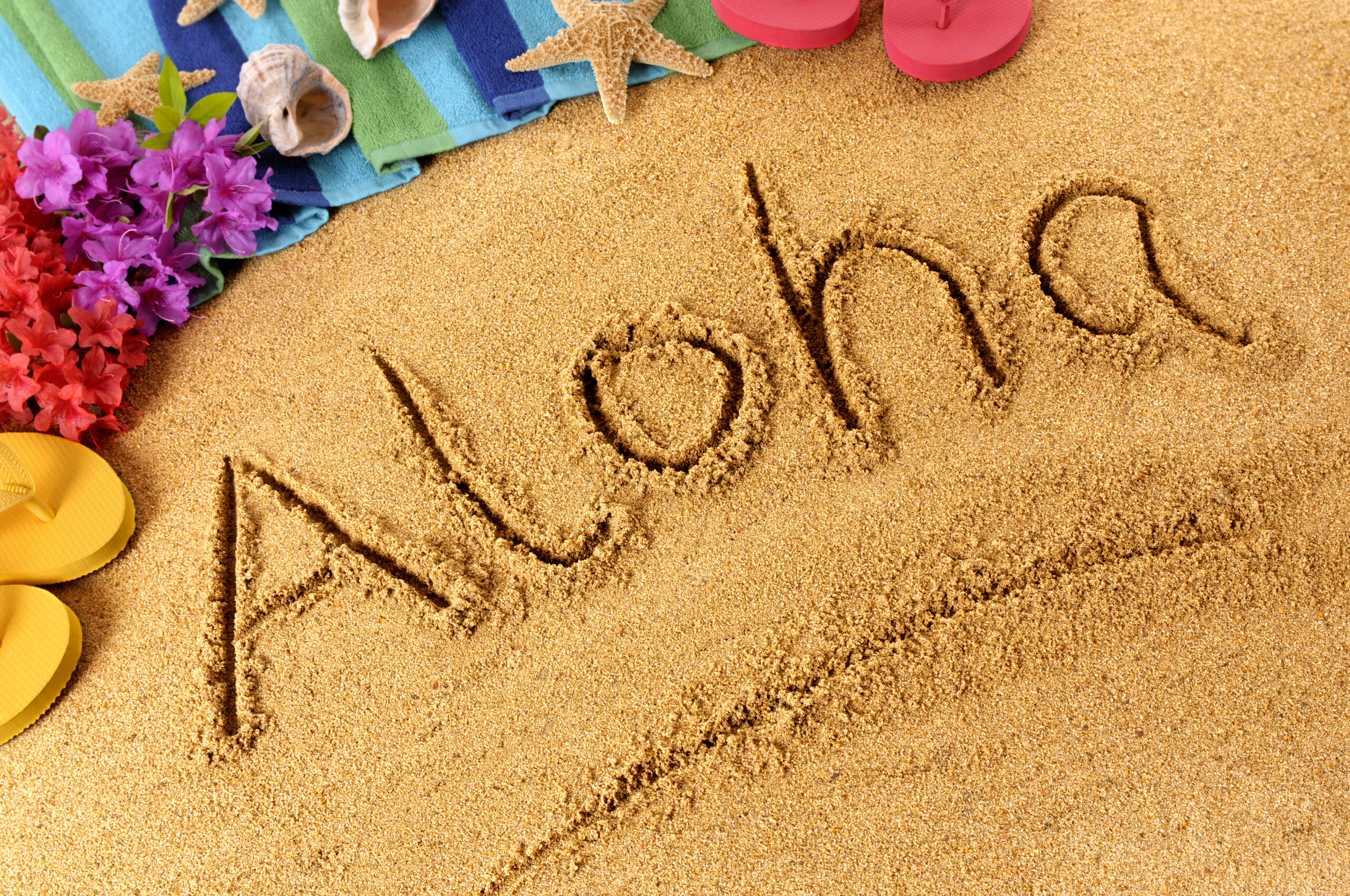 Aloha written in the sand surrounded by colorful flowers, beach towels, and flip-flops, symbolizing Hawaiian vacation experiences and tropical adventures.